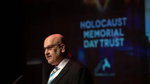 The image shows a bald man, dressed in a dark suit and wearing a white shirt with a yellow and blue striped tie, standing in front of a backdrop that reads 'Holocaust Memorial Day Trust.'