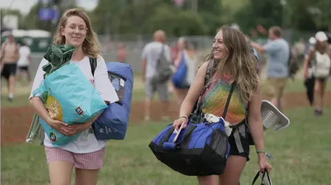 Girl smiles at friend whilst walking through a field carrying their luggage