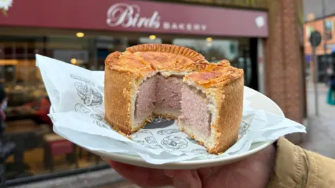 A large pork pie with a quarter cut out sits on a wrapper and plate in front of a bakery. The plate is being held be someone out of shot. The bakery has a large glass front and the maroon sign reads 'Birds Bakery.' There is a pavement and road sign beside it.
