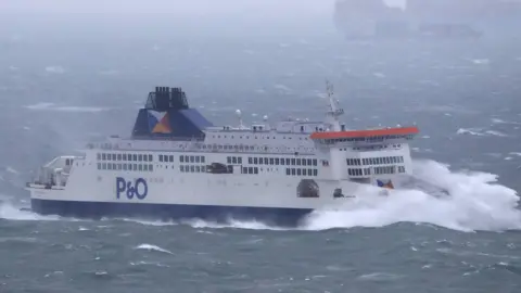 A blue and white P&O ferry in the sea. A large wave is crashing against the back. There are large waves in the water.
