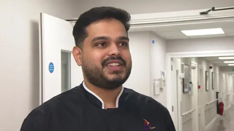 Mohan Sivashankar, a worker in a care home. He is standing in a care home corridor and wearing a navy blue shirt. He has short, black hair and a beard.