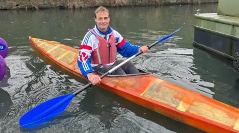 A Caucasian man sitting inside an orange kayak boat holding a black and blue paddle stick. The boat is in the canal. The man is wearing a Team GB jacket that is white, red and blue. He is also wearing a red life jacket. 