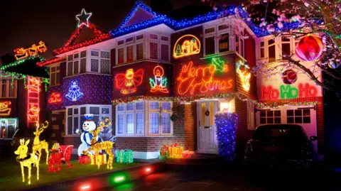 Getty Images Semi detached house pictured at night with various christmas light decorations including a full santas sleigh and reindeer on the front grass surrounded by light up presents and a huge 'merry christmas' sign lit up above the door.