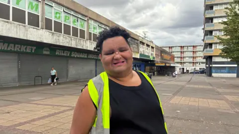 A young man stands smiling at the camera. He has short black curly hair and is wearing a black T-shirt and hi-vis jacket. Behind him are shops shut with shutters down and a paved square.