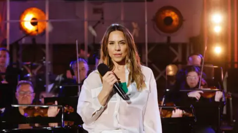 A woman, Melanie C, holding a mic, wearing a white shirt, with an orchestra behind her.