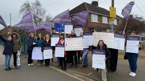 The photo shows a group of people smiling for the photo. They're each holding vairous signs and purple flags which read UNISON on them. 