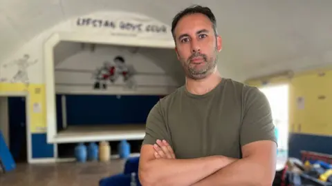 Stuart Woodward/BBC Jay Solder stands inside the Lifstan Boys Club building. He wears a dark green T-shirt, has dark short hair and a stubbly beard. He stands with his arms folded across his chest. Behind him is a stage with a boxing ring in the middle, and above the stage are the words "Lifstan Boys Club"