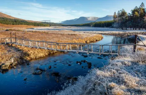 John Dewar Narrow bridge over a stream. One side of the bridge is covered in frost and the other is grass with sunlight shining on it.