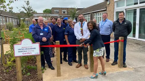 United Lincolnshire Hospitals NHS Trust Hospital trust director Sameedha Rich-Mahadkar cutting a red ribbon at the opening ceremony surrounded by staff