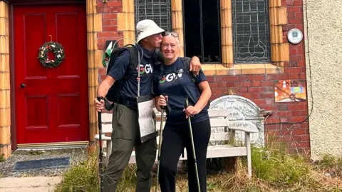 Andy Hockey John kisses Hayley on the forehead, with his hand around her shoulder. They hold walking sticks, and stand in front of a red brick house with a red door.
