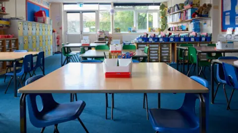 Tables and chairs arranged in a primary school classroom. At the back of the room is a window. There are no pupils or teachers present.