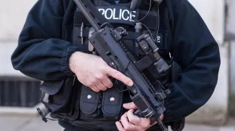A male police firearms officer pictured from shoulders to waist - he is dressed in all black with a bullet-proof vest and is holding a gun.
