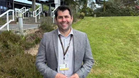 Leigh Frost is smiling as he stands in a garden with County Hall behind him. He has dark hair and brown eyes and is wearing a grey jacket and a grey and white striped shirt and a lanyard with his Cornwall Council identification on it.
