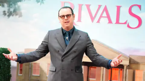Danny Dyer in a grey suit holding his arms out in front of Rivals branding at the premiere