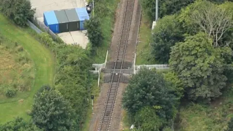 An aerial view of a railway line cutting through green woodland, with a fenced pedestrian crossing over the tracks and several coloured storage containers on a paved area nearby.