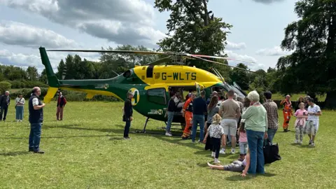 Wiltshire and Bath Air Ambulance The green and yellow Wiltshire and Bath Air Ambulance parked on a flat lawn with a queue of people standing next to it and trees in the background and cloudy sky.