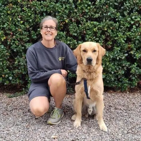 Bev Brenton Woman in dark jumper and shorts bent down next to a dog
