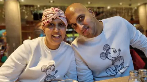 Priyanka Pandya-Bhatt Priyanka Pandya-Bhatt with her husband, Manoj Priyanka, sitting at a table, wearing white tops with Mickey Mouse on Priynaka, has a headscarf on and Manoy has a bald head. They are in a restaurant, with salt and pepper bottles on the table. 