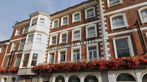 A google maps street view of The Cathedral Hotel in Salisbury. The building is red brick with white details. There is a row of hanging flowers across the width of the building. There are many windows and a sign reading "The Cathedral Hotel" in capital letters. 