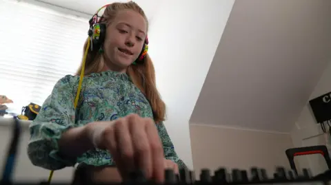 An action shot of a 10-year-old girl with long blonde hair twisting dials on a DJ deck in an attic room in a residential house.