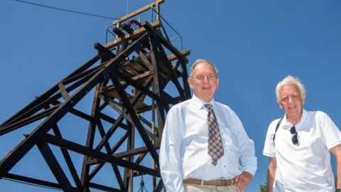 Volunteers Mike May and Roy Morton are standing in front of a 12m tall wooden structure which they helped to restore.
Mike is wearing a tie and a white shirt and Roy has a white polo shirt and has sunglasses attached to his shirt.