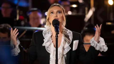 The singer Perrie standing onstage, singing into a microphone while looking upwards.