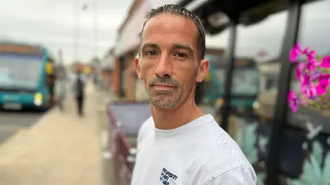 Stuart Woodward/BBC Matt Percy wears a white T-shirt and is standing in front of a shop. There is a bus in the background with people next to it. Matt has short dark hair and stubble.