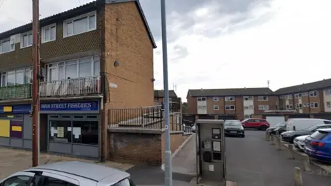 LDRS The location of the mural is a gable end of a brick building, in a residential area. There is a fish and chip shop in the building within a row of shops, and flats above. A phone box is in view as well as parked cars.