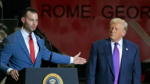Getty Images Republican candidate Clay Fuller, left, speaks behind a podium while Donald Trump stands off to his right, watching. Trump endorsed Fuller for Congress to replace retired Congresswoman Marjorie Taylor Greene.