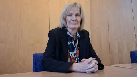 Susan McGill has blonde hair down to her shoulders. She is wearing a dark blue suit jacket over a floral pattern blouse. She is sitting in Stirling Council's chambers.