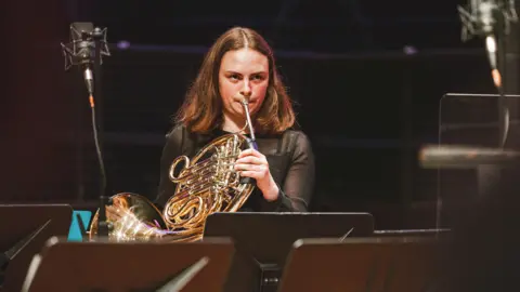 A woman in black formal wear plays the French horn on stage, surrounded by music stands for sheet music