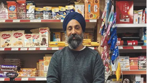 Southampton Forward A Sikh corner shop owner in a grey sweatshirt and blue turban, with a grey beard standing in front of shelves containing cakes and biscuits