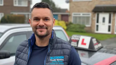 A dark-haired man with a dark beard, wearing a blue jumper and blue gilet, standing in front of two cars. One is silver, the other is a car used for learning to drive with a red and white L plate on the roof.