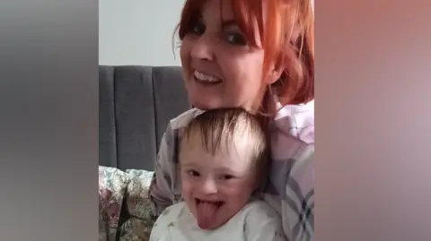Kirsty Haynes Kirsty Haynes sits smiling with her son Marco on her lap. She has bright red hair and a fringe and it is tied up. Marco is a toddler and is smiling and is sticking his tongue out.