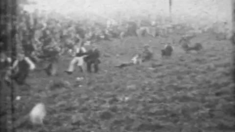 Unknown Black and white frame from a film showing a hazy pictures of a group of people running down a hill. There is a large white object in the right hand corner. 