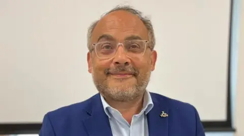Man with glasses, grey hair and beard, wearing a blue suit looks into the camera.