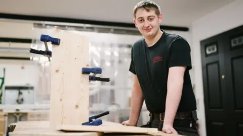 York Minster A young man stands in a joinery workshop. On the table in front of him is a piece of wood with clamps attached.