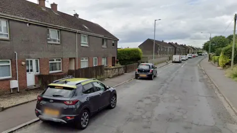 A residential street lined with terraced houses, showing several parked vehicles along the road, including a dark SUV in the foreground, with cloudy skies overhead.