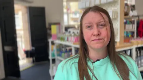 BBC/Nathan Turvey Kirsty Devine, who has brown long hair standing in her shop, there are shelves with items for sale on them and a door in the background.