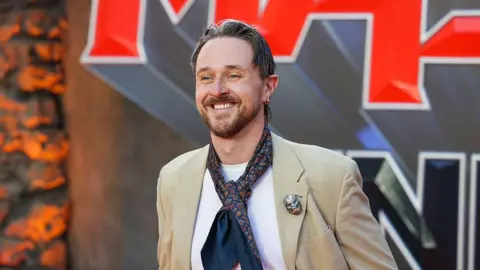 Getty Images Sam Wilson at a red carpet event. He is wearing a beige suit jacket and a tie put loosely around his neck, and a white t shirt. He has light brown hair and is smiling