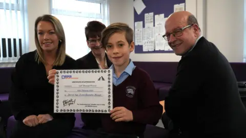 John Fairhall/BBC A young boy holds up a certificate from the RSPCA with three adults surrounding him. All of them are smiling at the camera.