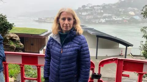 A woman wearing a blue puffer coat stood in front of a red plastic barrier fence, with water in the distance behind her. She has blonde hair.