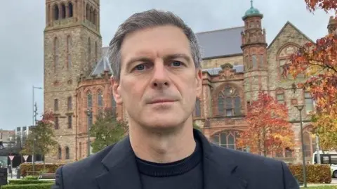 Shows a man in black jumper and black jacket with the Guildhall in Derry in the background