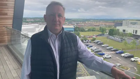BBC/Laura Jones A man with short brown hair is wearing a pale shirt with small checks and a body warmer. He is standing on a balcony which overlooks a car park and industrial units