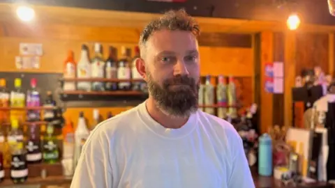 Matthew Nichol, who wears a white t-shirt and has a beard, looks at the camera inside The Flying Dutchman pub