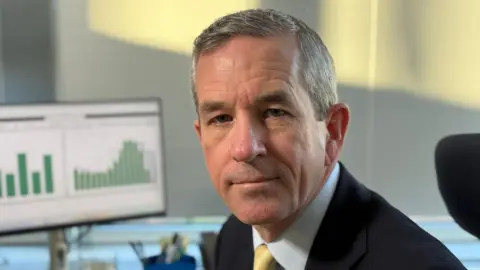 Steve Hubbard/BBC Neill Moloney wearing a black suit jacket, pale blue shirt and golden tie. He has short grey hair and looks serious while sitting at a desk, which has a computer monitor on it.