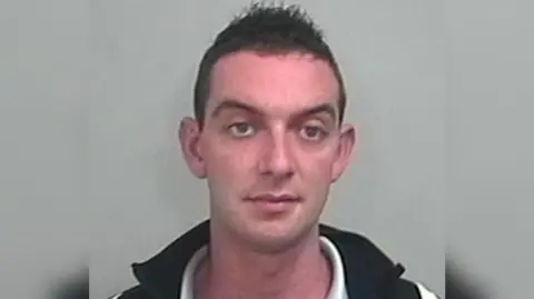 A man with short dark hair looking towards the camera wearing a black vest over a white shirt