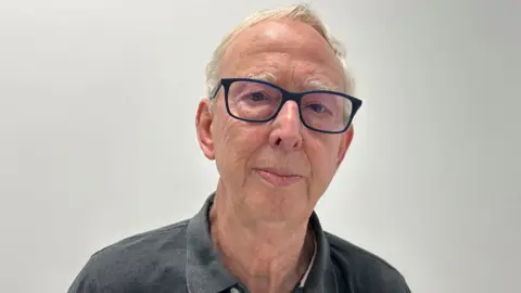 BBC / Gavin Kermack An elderly man with short white hair and dark-rimmed glasses, wearing a grey polo shirt, stands in front of an off-white wall.