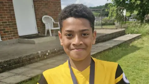 Nicky Ajanaku A young boy wearing a yellow and black football shirt looking straight at the camera and smiling. He is stood outside on grass in front of a building. 