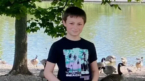 Family Leo Ross, a young boy with brown hair, smiling at the camera. He is in a park. There is a body of water and geese behind him. He is wearing a black top with a colourful blue and purple pattern on. 